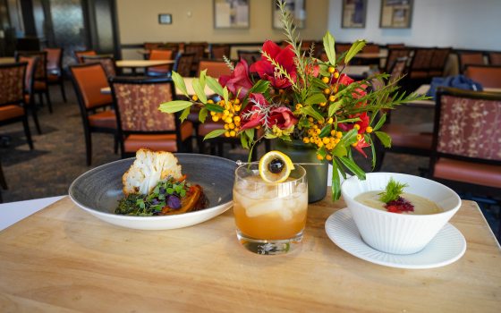 a plate of food, drink, bowl of soup and a vase of flowers on a wood table